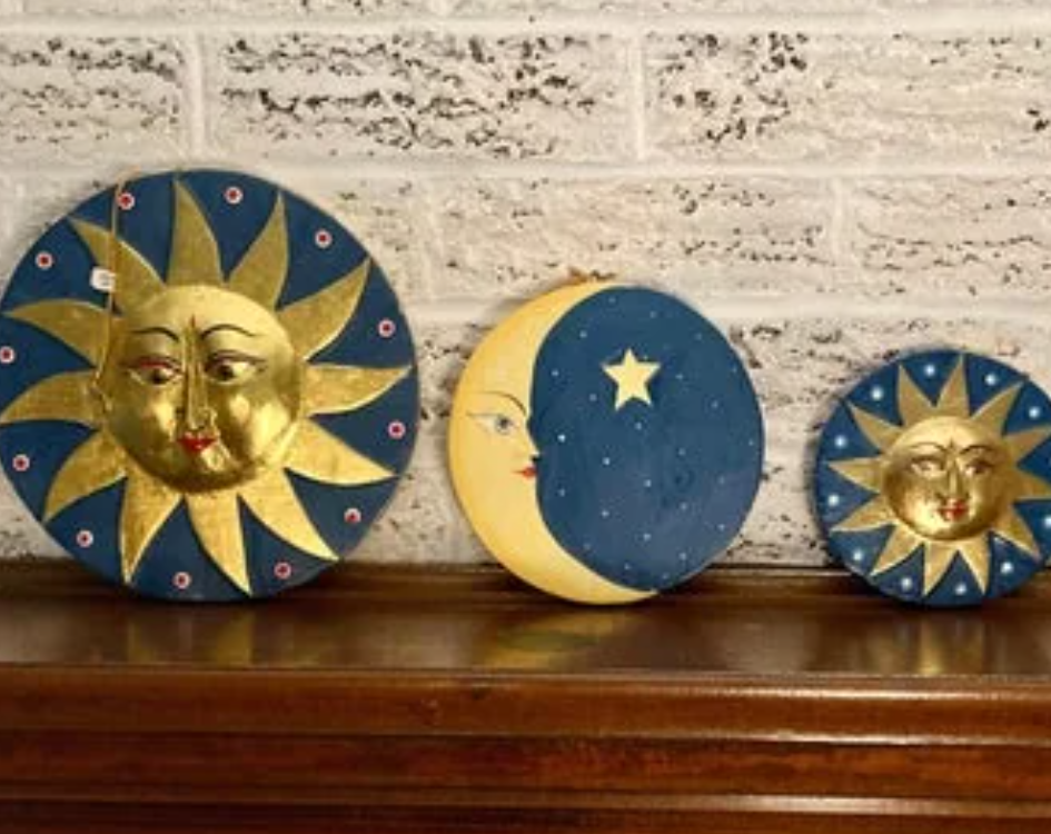 A set of three decorative wall plaques with celestial themes, popular in the 90s and 2000s. The plaques feature a golden sun with a smiling face and a blue background with stars, a crescent moon with a serene face and a single star, and a smaller golden sun with a blue background. The plaques are displayed against a white brick wall on a wooden mantel.