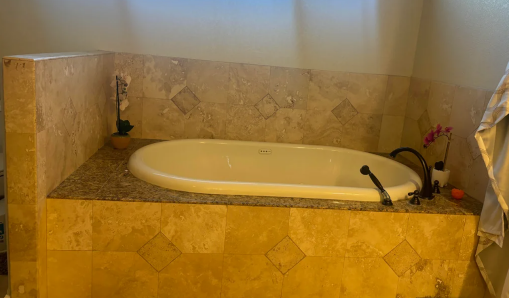 
A luxurious bathroom featuring a built-in bathtub surrounded by beige marble tiles, typical of the 2000s design. The bathtub area includes a granite ledge, black fixtures, and decorative orchids placed on either side. The walls and ledge are accented with diamond-patterned tiles, enhancing the elegant and serene atmosphere of the space.