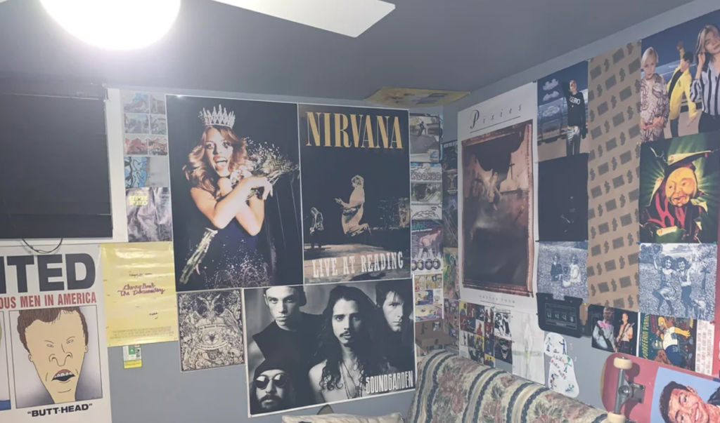 A room with various music and pop culture posters on the walls. Prominent posters include Nirvana, Mariah Carey holding flowers and wearing a crown, Pixies, and a Beavis and Butt-Head poster. The walls are decorated with a mix of band posters, album covers, and other visual art.