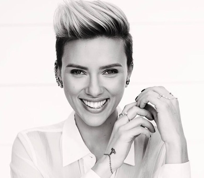 A black-and-white portrait of Scarlett Johansson smiling with short, styled hair. She is wearing a light-colored shirt and multiple rings, with a tattoo visible on her wrist.