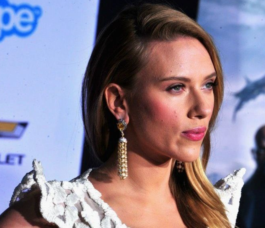 Scarlett Johansson in a white lace dress with long, dangling earrings, looking off to the side at an event. The background features a partially visible Chevrolet logo and a blue-and-white backdrop.