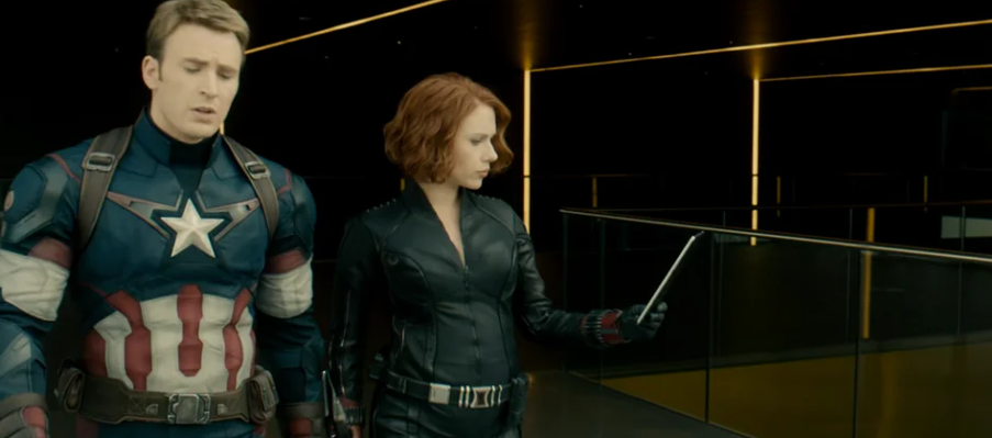 A scene from a movie featuring Chris Evans as Captain America in his superhero costume and Scarlett Johansson as Black Widow in her black tactical suit. They are walking together in a modern, dimly lit setting, with Black Widow holding a tablet. Johansson is visibly pregnant.