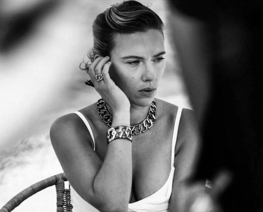 A black-and-white photo of Scarlett Johansson wearing a chunky chain necklace and bracelet, with her hand touching her face. She is dressed in a sleeveless top, looking contemplative while sitting on a chair.