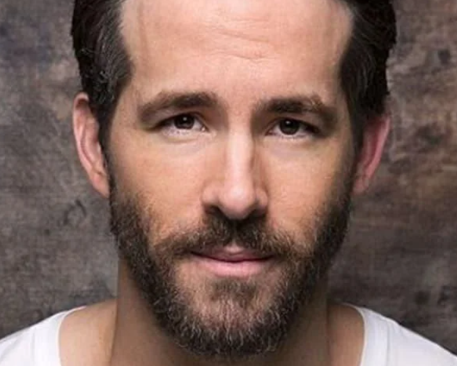 Close-up of Ryan Reynolds with a short beard and mustache, wearing a white shirt and looking directly into the camera with a slight smile. The background is a textured, neutral-toned backdrop.