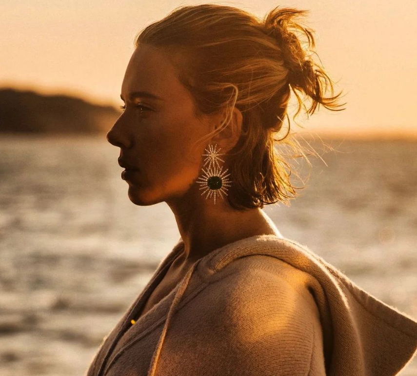 Scarlett Johansson in profile, bathed in golden sunlight near a body of water. She is wearing a cozy sweater and statement earrings with a sunburst design. Her hair is tied back in a loose, casual style.