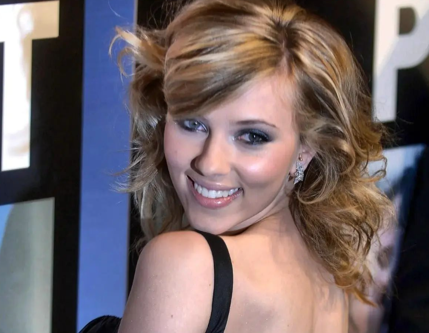 Scarlett Johansson smiling over her shoulder at a red carpet event. She has wavy blonde hair, wears a black dress with bare shoulders, and has sparkling earrings. The background includes part of a poster or banner.
