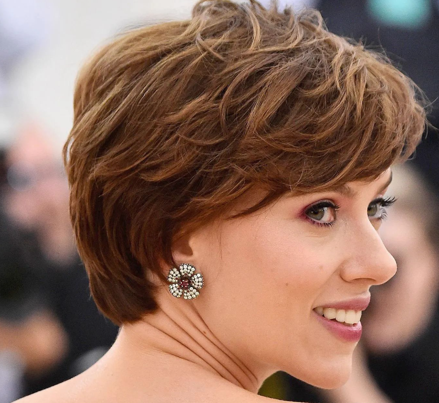 Scarlett Johansson at an event, smiling over her shoulder. She has short, tousled brown hair and is wearing floral-shaped earrings. The background is blurred, focusing attention on her face and hairstyle.