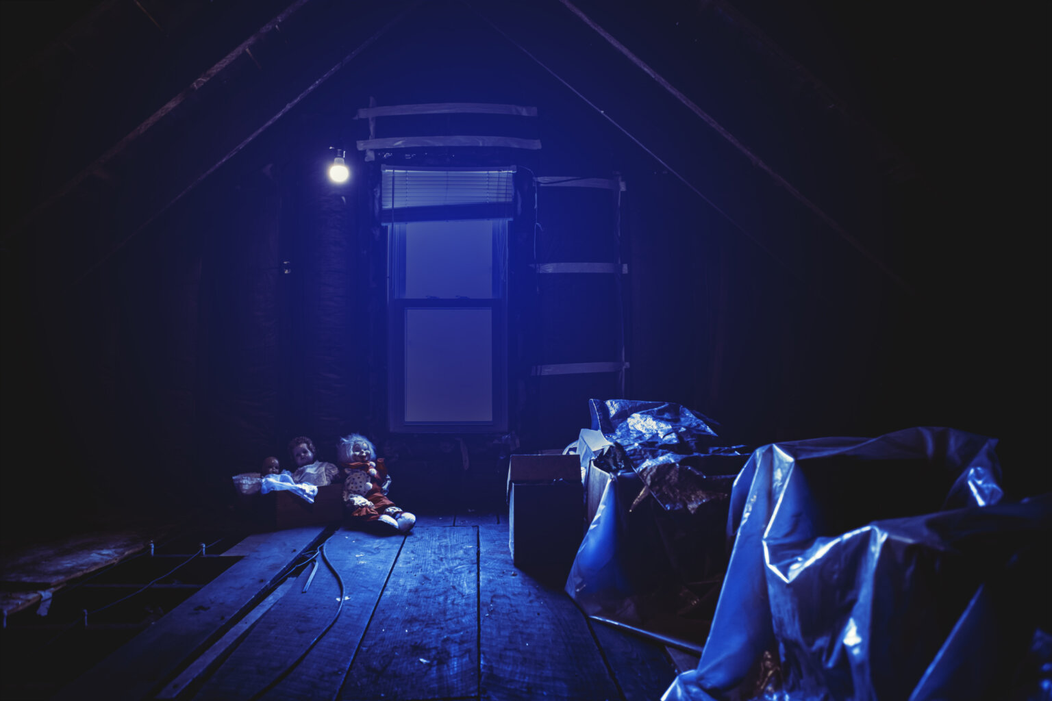 A dimly lit attic with exposed wooden beams, illuminated by a single blue light. Two old dolls sit eerily on the floor near a window, adding a spooky atmosphere. The attic is cluttered with plastic-covered items and various objects scattered around.