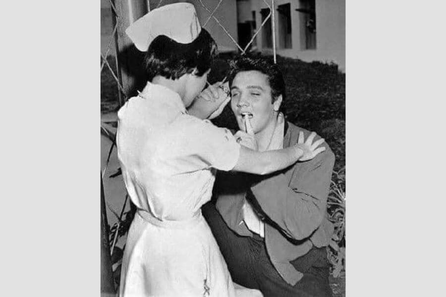 A young Elvis in his twenties pointing to his own mouth while being treated by a nurse