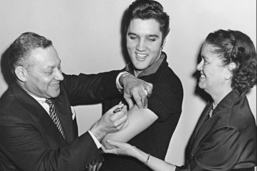 A man applies a vaccine to Elvis Presley's arm while a woman holds his arm.