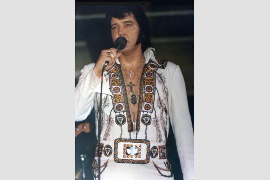 Elvis Presley holding a microphone on stage while wearing a cross, a chai symbols and a star of david around his neck. 