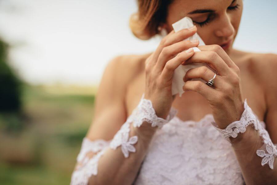 Beautiful bride in white dress weeps tears of happiness on the wedding day. Emotional woman in wedding gown wipes the tears with tissue paper.