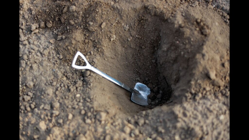 A metal shovel in an earthen pit, surrounded by soil and dirty earth. Digging a hole.