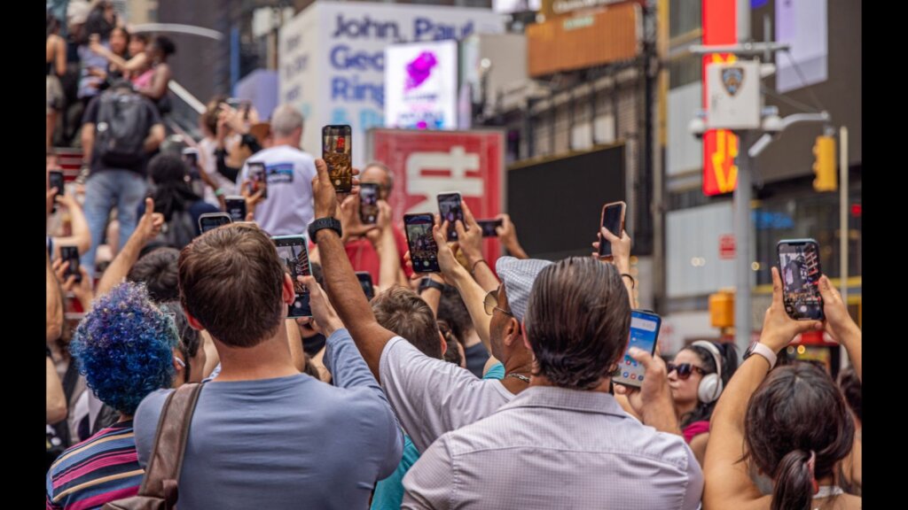 Large crowd of people taking pictures of the neon signs and tall buildings with their mobile phones.