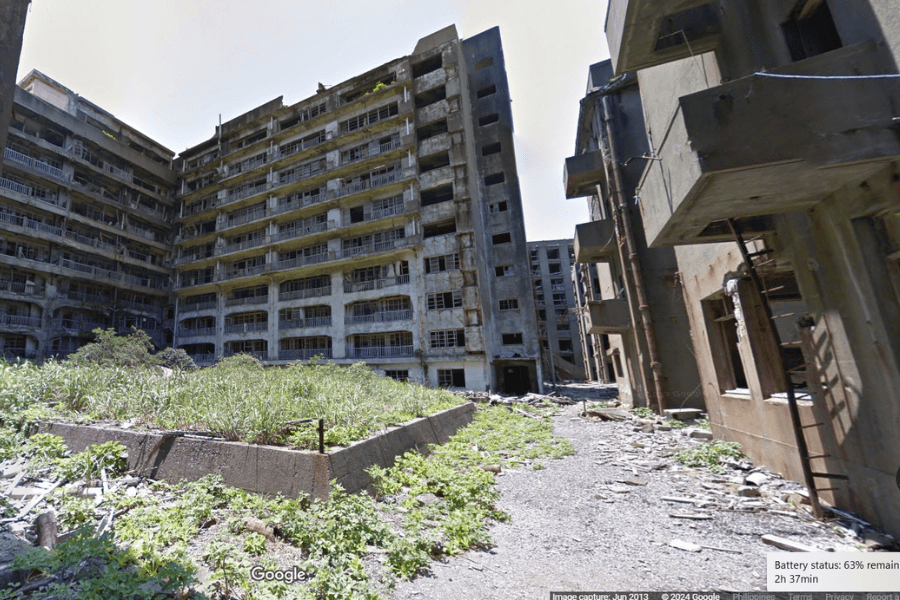 This image shows an abandoned, deteriorating multi-story building complex, with overgrown vegetation and rubble scattered across the ground, suggesting a desolate and neglected location.