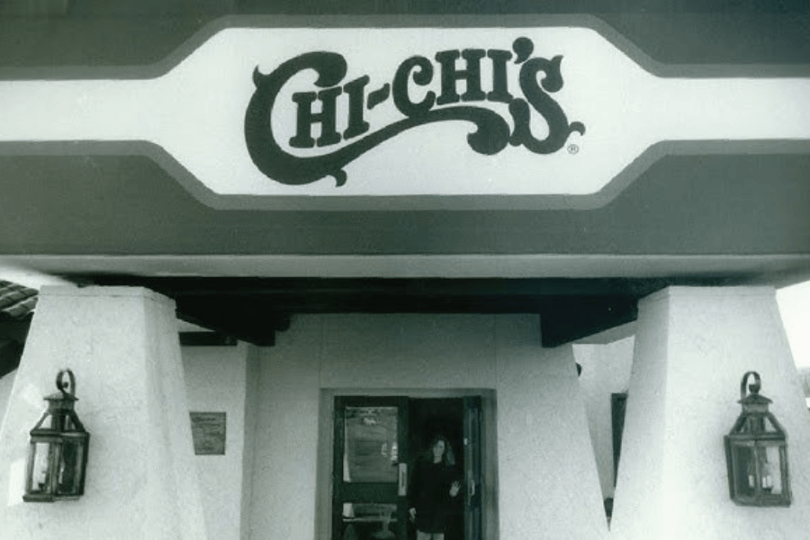 The photo shows the entrance of a Chi-Chi’s restaurant, featuring a large sign with the restaurant's logo above the doorway and a classic architectural design with lantern-style lights on either side.