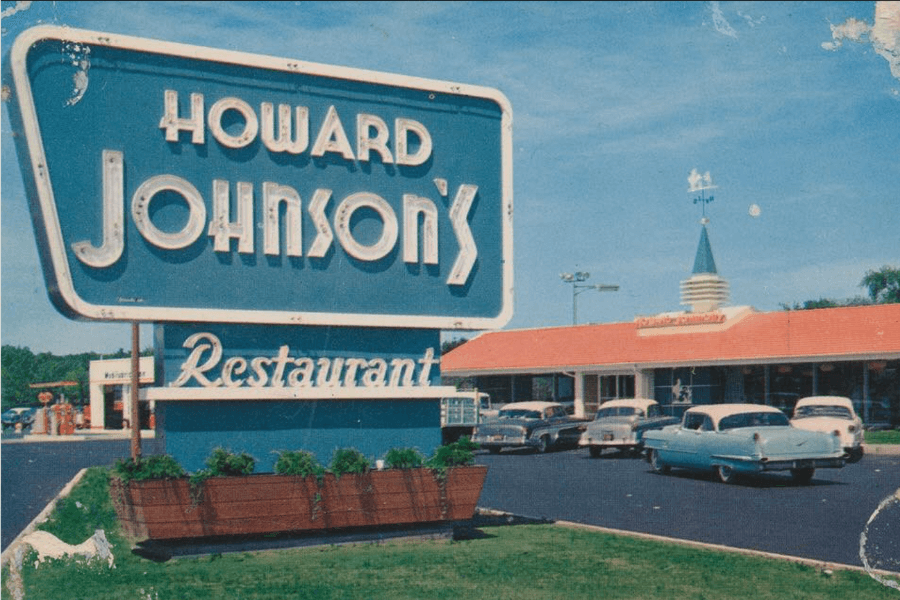 The image shows a classic Howard Johnson’s restaurant with its iconic blue sign and retro lettering, surrounded by vintage cars in the parking lot, evoking a nostalgic snapshot of mid-20th-century Americana.