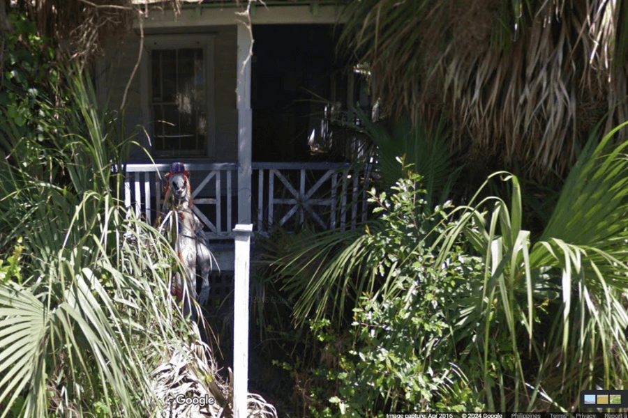 This image shows the front porch of a house partially obscured by dense, overgrown foliage, with a mannequin or statue resembling a creepy figure in a top hat standing near the entrance. 