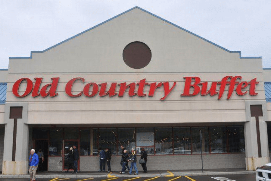 The image shows the entrance of an Old Country Buffet restaurant, with its bold red lettering on the facade. People are seen entering and exiting, evoking memories of hearty meals and endless buffet options.