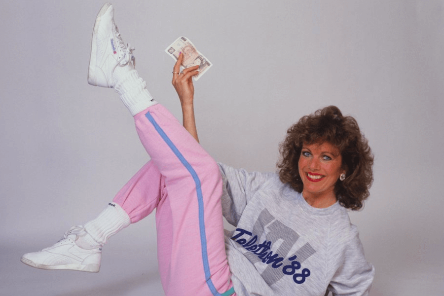 The photo shows a woman in '80s-style fitness attire—pink sweatpants, white sneakers, and a sweatshirt—cheerfully posing with one leg raised and holding a banknote in her hand, radiating a playful and energetic vibe.