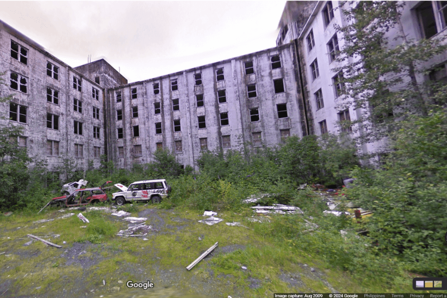 This image shows an abandoned, dilapidated multi-story building with numerous broken windows and weathered walls. In the foreground, there are rusted and damaged vehicles surrounded by overgrown vegetation and scattered debris, enhancing the sense of neglect and desolation. 