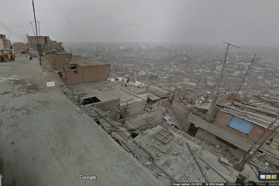 This image shows a hillside neighborhood with densely packed, improvised buildings overlooking a sprawling urban landscape under a gray, overcast sky. It shows an elevated road around residential areas and the road has no protective barricades.