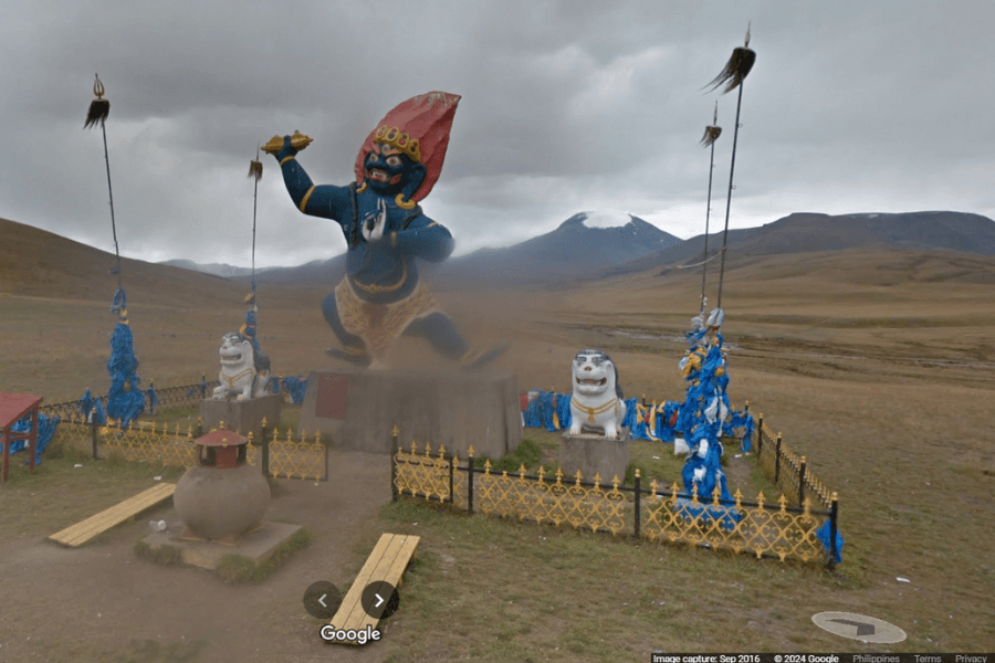 This image depicts a large, colorful statue of a fierce, mythological figure standing in an open field surrounded by mountainous terrain under a cloudy sky. The statue, with its striking red headdress and expressive pose, appears to represent a deity or protective spirit, possibly from a Buddhist or Mongolian cultural context. 