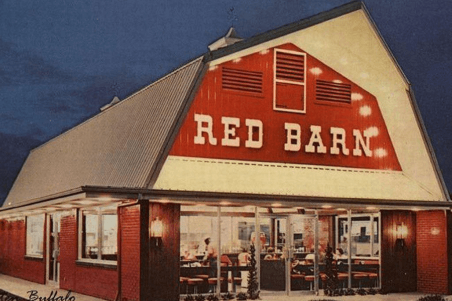 The image shows a Red Barn restaurant with its iconic barn-shaped design, illuminated signage, and inviting windows that reveal a cozy interior. Its nostalgic architecture evokes memories of classic family dining experiences.