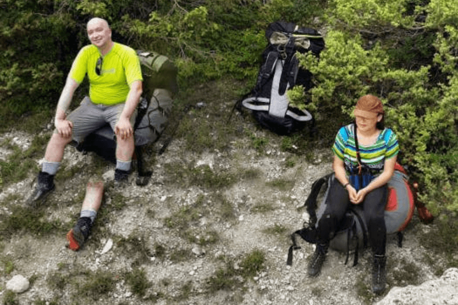 This image shows two hikers taking a break on a rocky ground surrounded by bushes. The man on the left is smiling and wearing a bright green shirt, sitting with his legs seems like amputated because of the glitch, while the woman on the right, wearing a striped shirt and a cap, sits on her backpack looking slightly away.