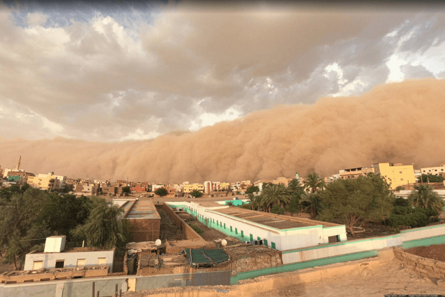 This image shows an enormous wall of dust from an approaching sandstorm engulfing a town with various buildings and greenery. The massive cloud of sand looms over the cityscape, creating an intense and dramatic contrast between the clear foreground and the dense, reddish-brown dust behind it. 