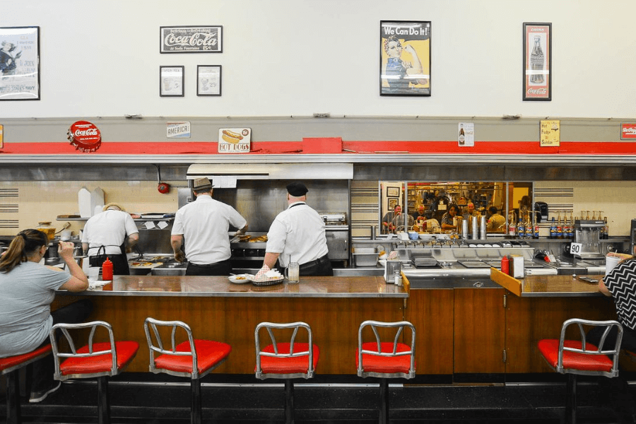 The image shows a classic diner with red swivel stools at the counter, where chefs in white uniforms prepare meals on a grill, evoking a nostalgic, retro dining atmosphere.