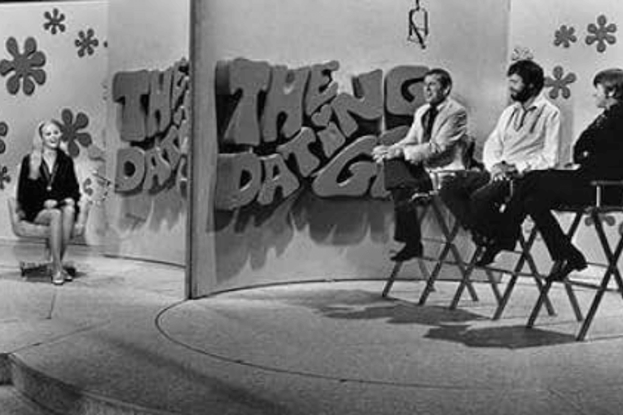 The black-and-white image is a scene from The Dating Game, a popular television show from the 1960s and 1970s.

Left Side: A female contestant sits on a chair, facing away from the three male contestants. She is likely the "bachelorette," who will ask questions to the bachelors without seeing them, as was the format of the show.

Right Side: Three male contestants sit on high chairs, separated from the female contestant by a decorative divider that prominently displays the show's logo, "The Dating Game," in bold, playful lettering. Each contestant appears relaxed, waiting to answer questions in hopes of being selected for a date.