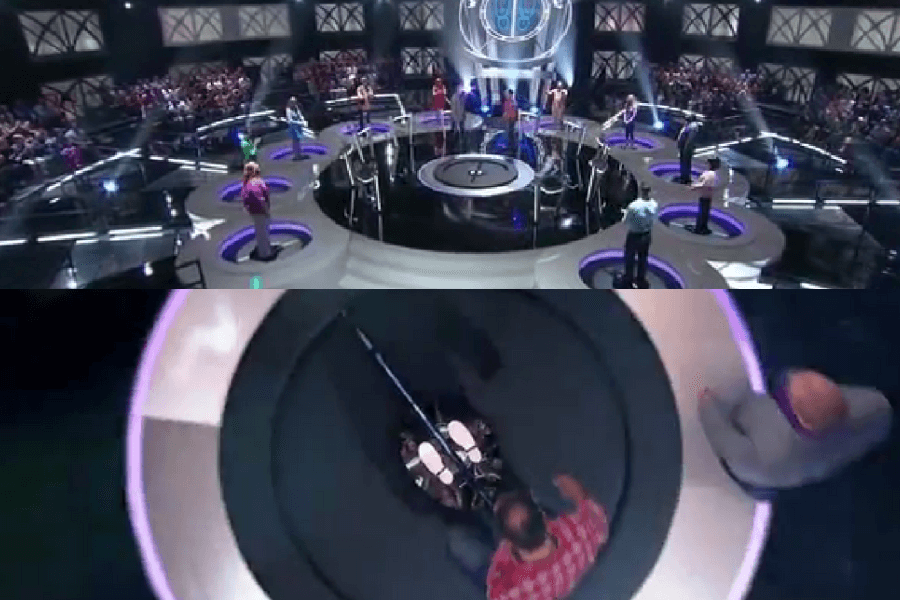 The image shows contestants on the game show Who’s Still Standing?, where each participant stands on a trapdoor while answering trivia questions. If they get a question wrong, the trapdoor opens, and they dramatically drop through the floor. 