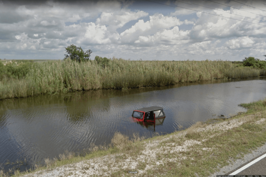 This image shows a partially submerged red vehicle in a body of water, possibly a canal or roadside ditch, with only the roof and windows visible above the surface. The surrounding area is grassy with tall reeds, and a road runs alongside the water.