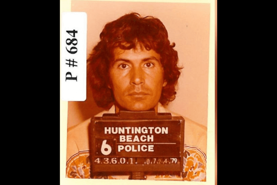 The image is a mugshot of Rodney Alcala taken by the Huntington Beach Police. Alcala is shown with a serious expression, facing directly toward the camera. The placard in front of him reads "Huntington Beach Police" along with the arrest date, "07-24-79," and his case number. He has curly, medium-length hair and wears a patterned shirt, which adds to the 1970s aesthetic.