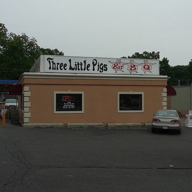 A small barbecue restaurant named "Three Little Pigs Bar-B-Q" with a beige exterior and a large white sign on the roof featuring cartoon pigs and stylized text. A neon "OPEN" sign is visible in one of the windows, and a single car is parked in front of the building. The restaurant has a simple, unassuming design with red awnings on the sides.