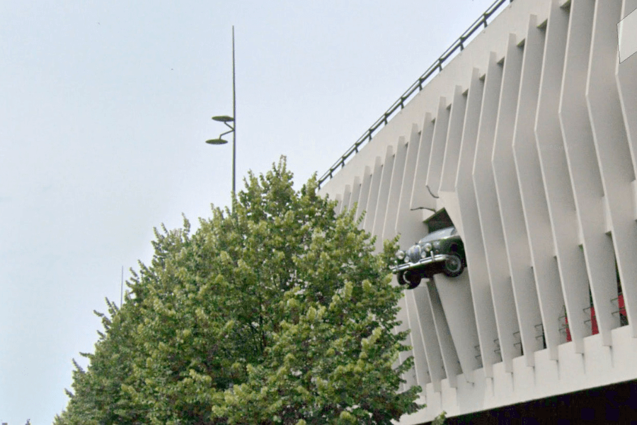 This image shows a car seemingly embedded in the side of a modern building, as though it has burst through the wall. The vehicle is partially protruding from the structure, creating an optical illusion or artistic installation. The design suggests that this is an intentional display rather than an actual accident.