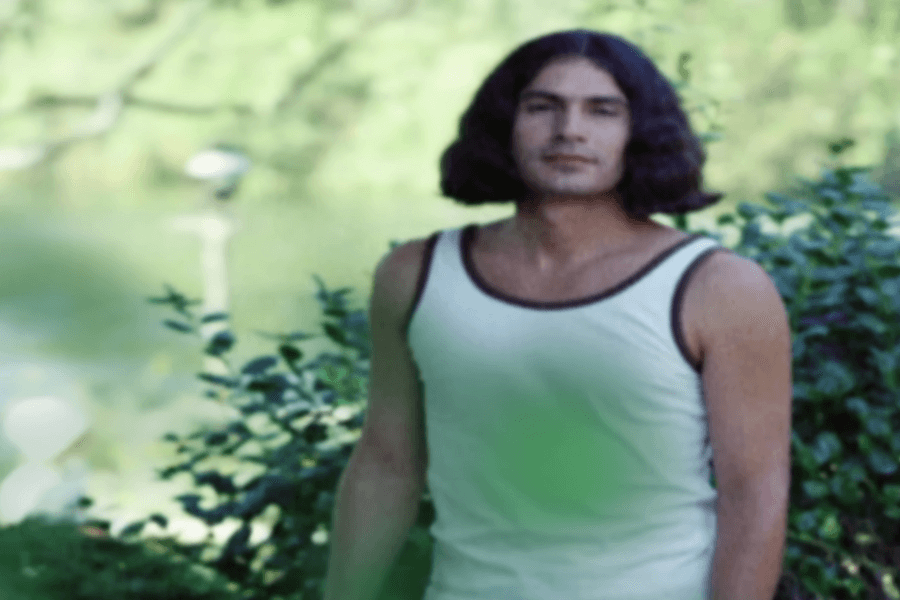 The image is a photograph of Rodney Alcala, taken outdoors. He is dressed casually in a sleeveless tank top with a dark trim, standing in front of lush greenery. Alcala has shoulder-length hair and a neutral expression on his face.