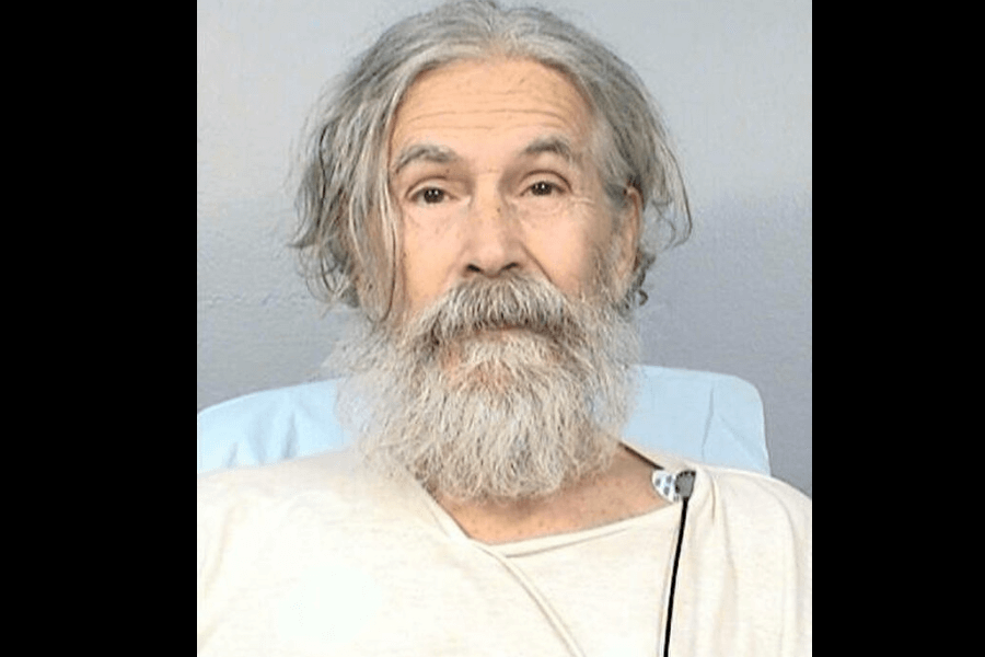 The image is a recent mugshot of Rodney Alcala in his later years, showing him with a long, gray beard and unkempt gray hair. He appears aged, with deep lines on his face, and is wearing a hospital gown, indicating that the photo may have been taken during a period of illness or confinement in a medical facility. His expression is calm, almost blank.