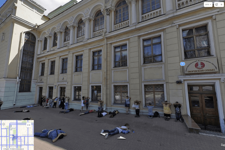 This image shows a group of people lined up against the wall of a large building with their hands raised or resting on their heads, while others lie on the ground nearby. 