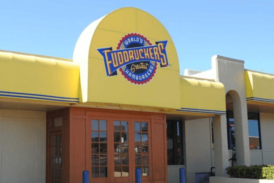 The image showcases the bright yellow exterior of a Fuddruckers restaurant, featuring its iconic logo that reads "World's Greatest Hamburgers," evoking memories of custom-made burgers and fresh, bakery-style buns.