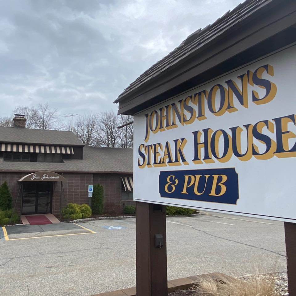 Johnston’s Steak House & Pub, a rustic restaurant with a brown exterior, striped awnings, and a chimney, sits under a cloudy sky. A large wooden sign in the foreground displays the restaurant’s name in bold gold and navy lettering. The parking lot is mostly empty, with a handicap space visible near the entrance.