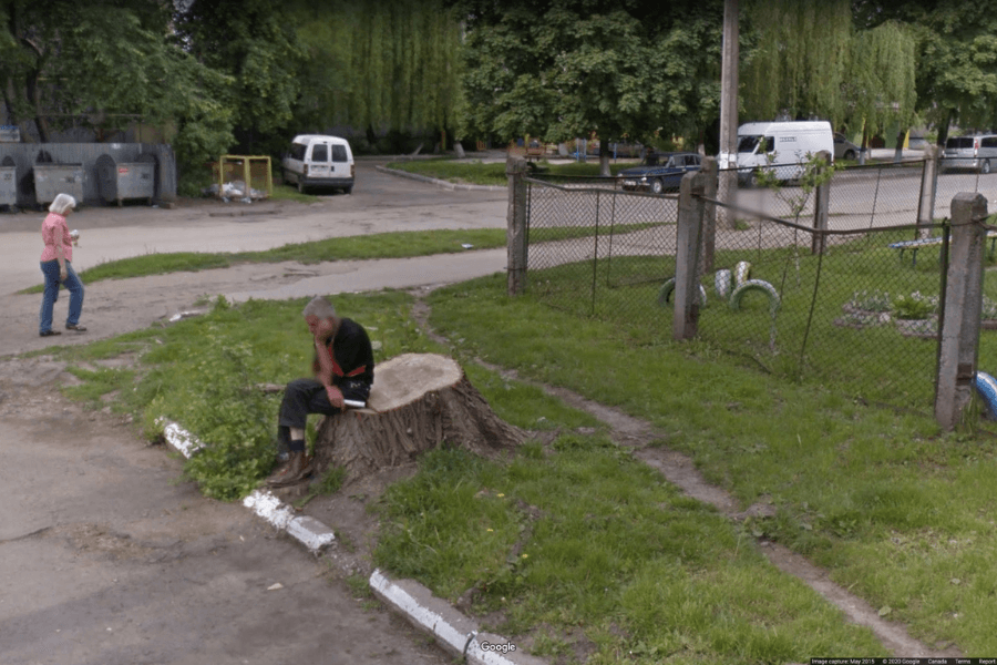 This image shows an older man sitting on a large tree stump in a small, grassy area near a fenced section and a few parked vehicles. The man appears to be holding his head in his hand, suggesting a contemplative or tired posture. 