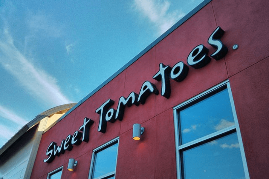 The image features the exterior of a Sweet Tomatoes restaurant, with its distinctive cursive logo set against a vibrant red wall, symbolizing its reputation for fresh salads and wholesome buffet options.