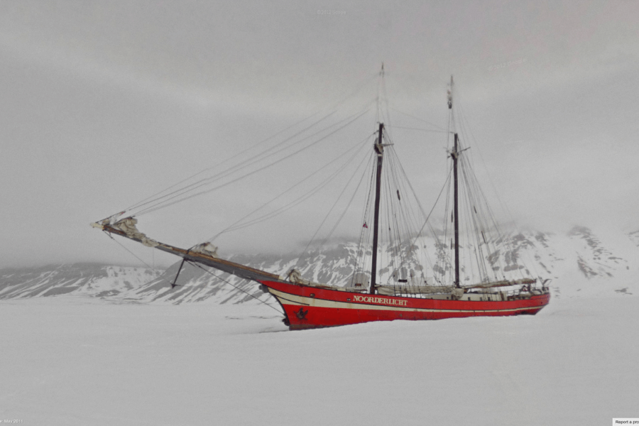 This image shows a striking red sailing ship named Noorderlicht trapped in a frozen, snowy landscape with snow-covered mountains in the background. The ship appears isolated, surrounded by ice and snow, creating a stark contrast against the monochromatic surroundings. 