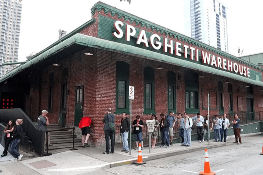 This image showcases the Spaghetti Warehouse, a vintage brick building with a green awning and bold signage. A line of people gathers outside, reflecting the restaurant’s popularity and nostalgic charm.