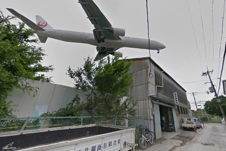 This image captures a low-flying airplane passing directly over an industrial area, with a warehouse and narrow street below. The aircraft, bearing the Japan Airlines (JAL) logo, appears very close to the buildings, creating a dramatic and intense perspective as if it’s about to land or take off nearby. 