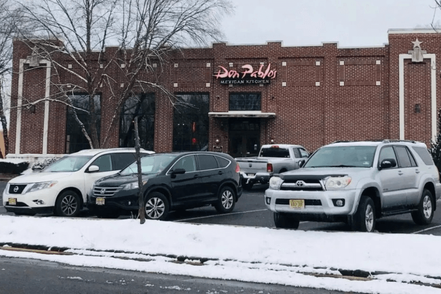 This image showcases the exterior of a Don Pablo’s Mexican Kitchen, a red-brick building with a bold neon sign, surrounded by snow-covered trees and parked cars, evoking memories of Tex-Mex dining experiences.