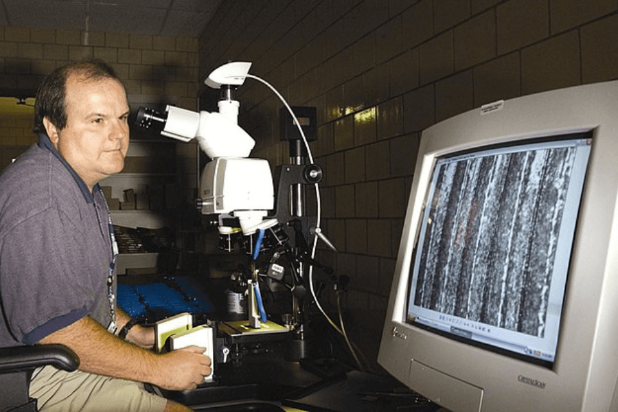 This image shows a forensic scientist or technician working with a high-powered microscope connected to a computer monitor displaying a magnified view of a sample. The individual appears focused on examining the sample, likely as part of forensic analysis or research.