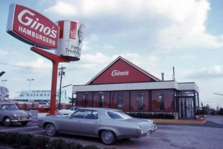 A vintage Little Tavern Shop restaurant with its signature green awning and pyramid-shaped roof. The awning displays the restaurant's name along with menu items like "Sausage & Eggs," "Cheeseburgers," "Burgers by the Bag," and "Hot Dogs." The small building has large glass windows, and the parking lot in front has a few yellow barriers.